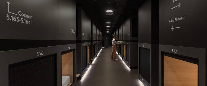 A dim corridor lined with stacked sleeping capsules, some open and lit from inside.