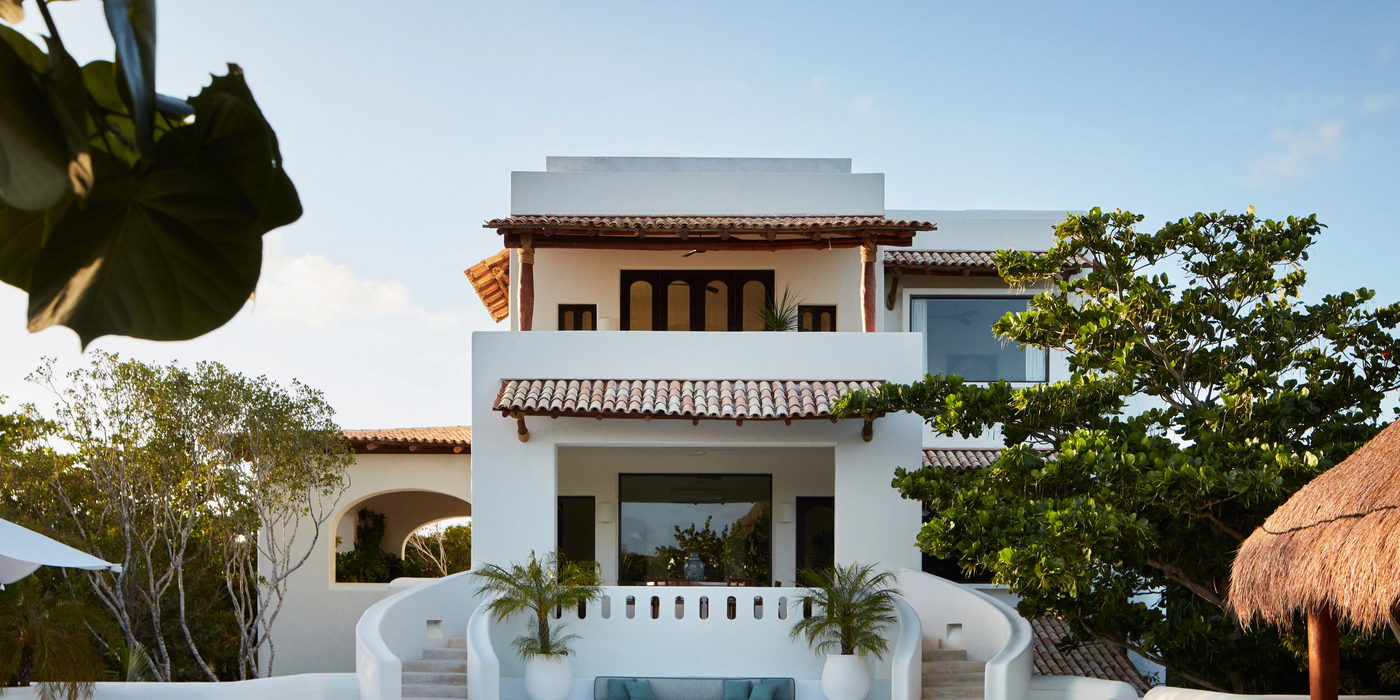 A white two-storey house with a curved double stair, set among palms.