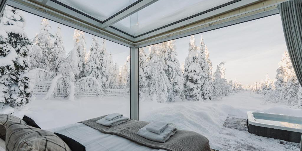A bed facing a glass wall and snow-covered trees, with a hot tub on the deck outside.
