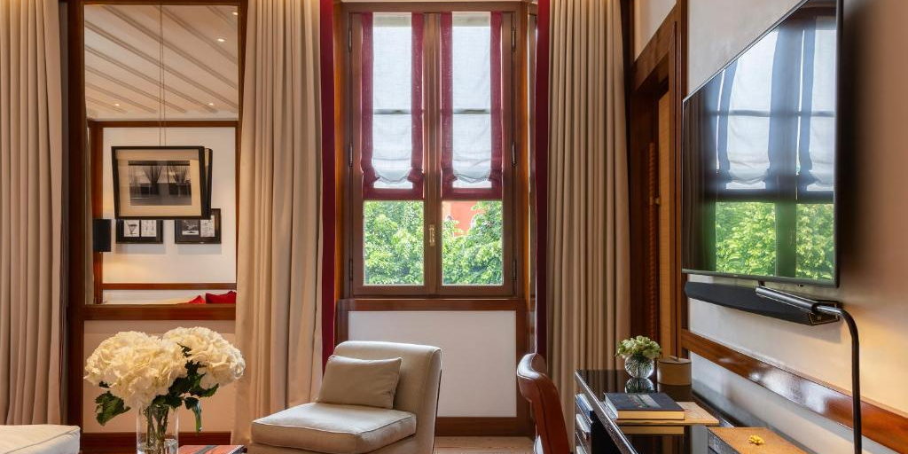 A hotel sitting room with a window, a cream armchair and a desk.